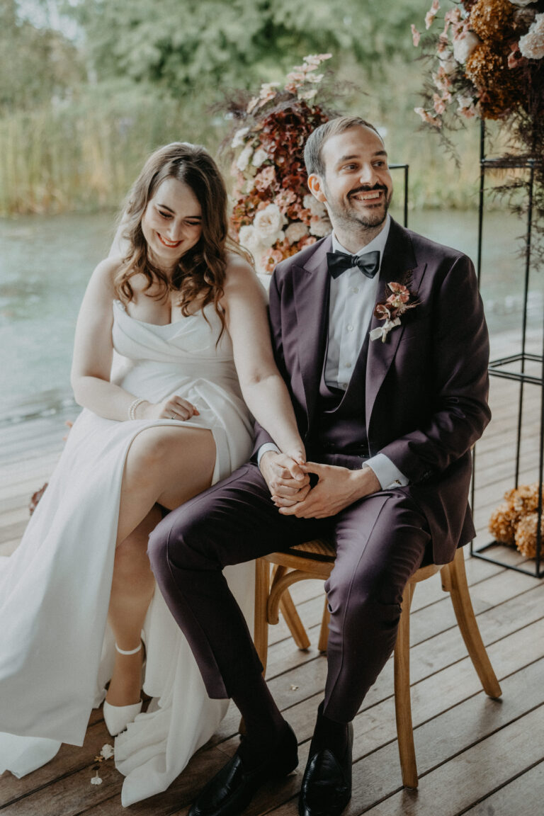 Braut und Bräutigam sitzen händchenhaltend und lächelnd, umgeben von Blumen auf einer Holzterrasse am Wasser - ein idyllischer Moment, perfekt für die Hochzeitsplanung in der Schweiz oder einen romantischen Italien-Urlaub.