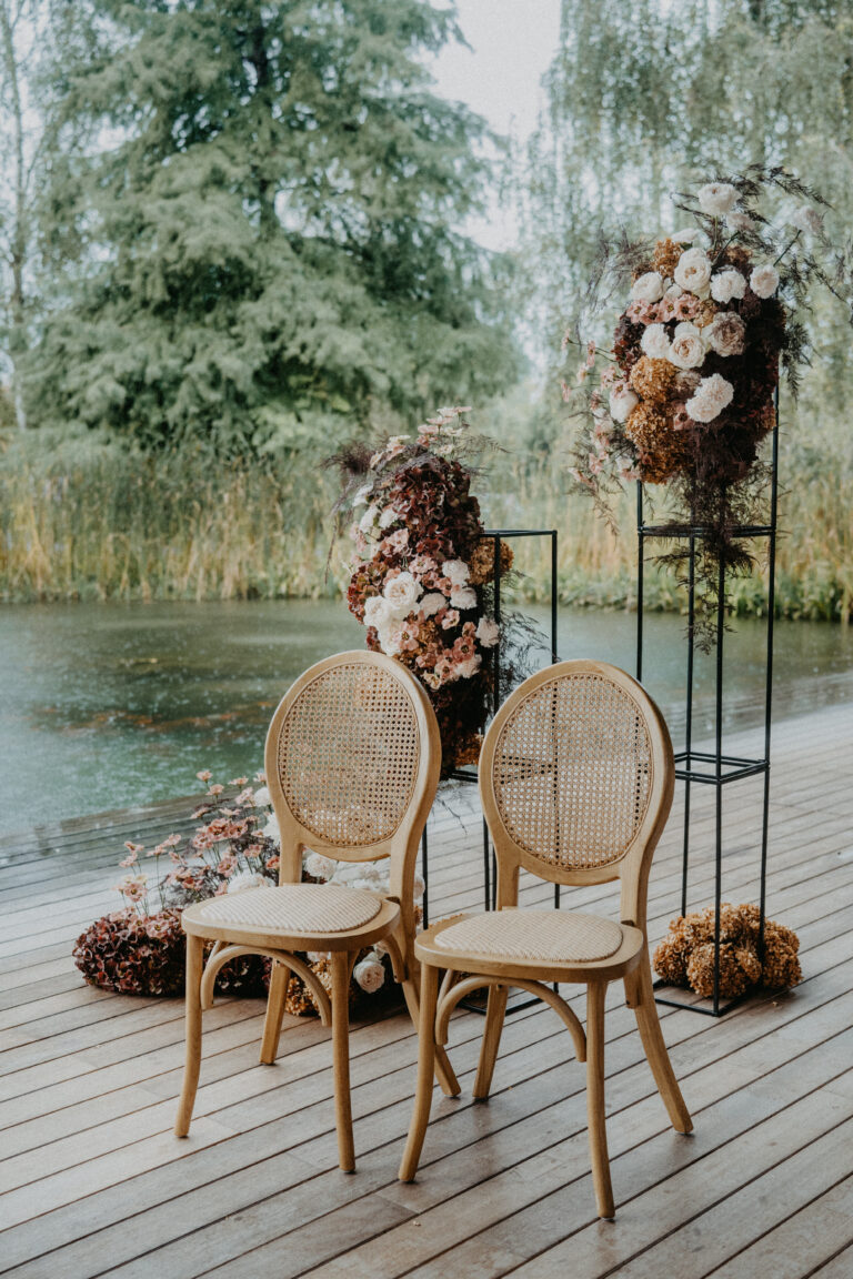 Zwei Holzstühle auf einer Terrasse an einem Teich, mit Blumenarrangements auf Metallständern im Hintergrund - eine traumhafte Kulisse, die jeder Hochzeitsplaner in Italien für eine intime Hochzeit lieben würde.