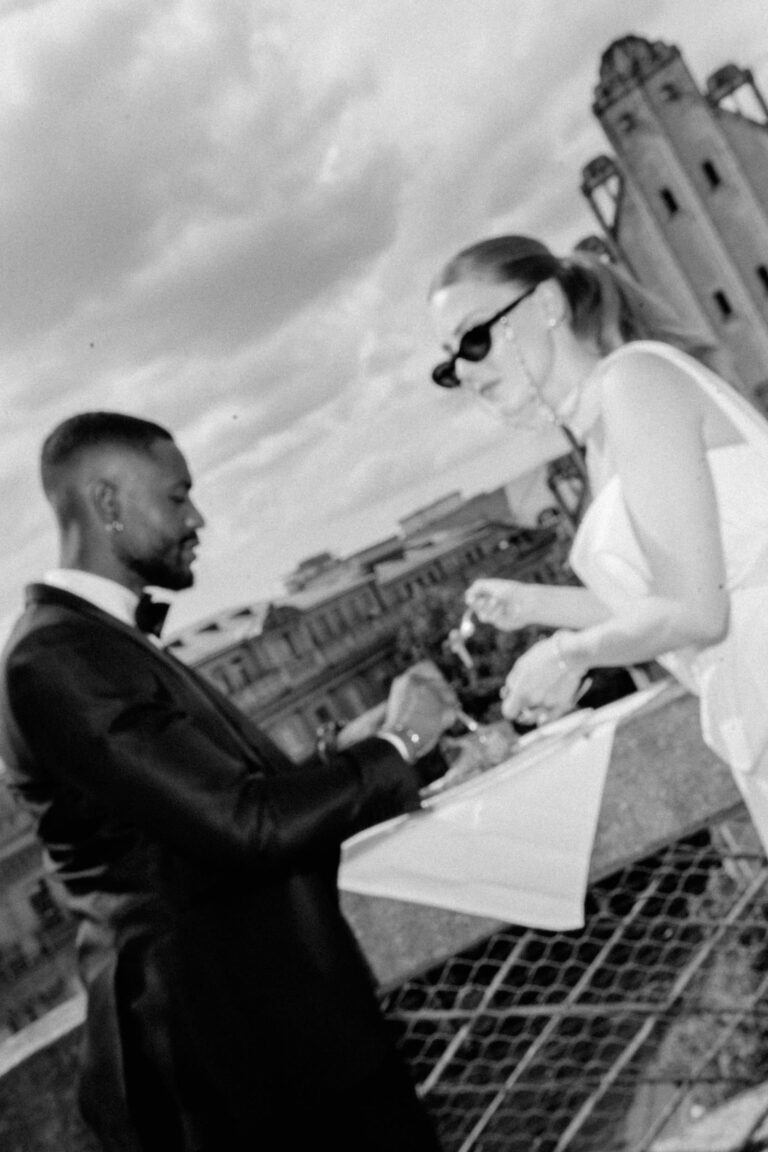 Ein Mann im Anzug und eine Frau mit Sonnenbrille, die vielleicht Einzelheiten mit ihrem Hochzeitsplaner besprechen, speisen an einem Tisch im Freien mit den Gebäuden der Stadt im Hintergrund.