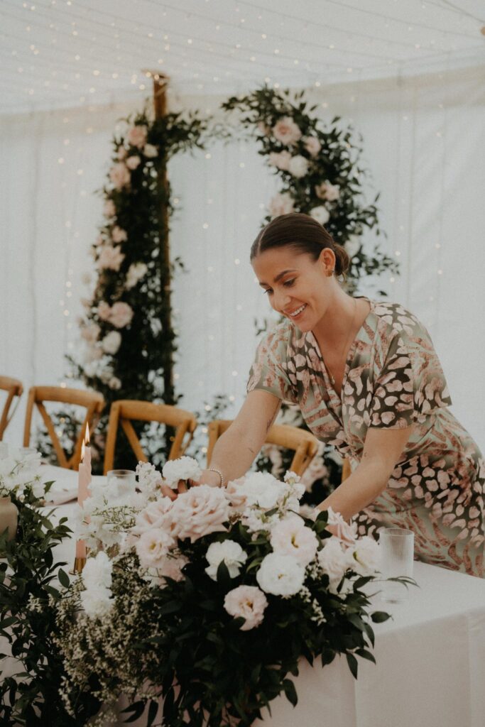Eine Frau arrangiert Blumen auf einem geschmückten Tisch bei einer Hochzeit oder Veranstaltung in Italien, mit einem schönen Blumenbogen im Hintergrund - die perfekte Inspiration für die Hochzeitsplanung.