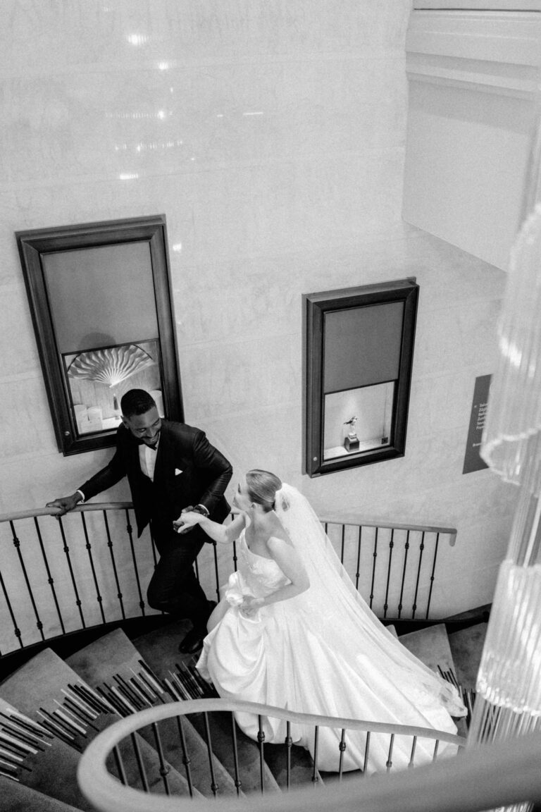 Eine Braut und ein Bräutigam gehen eine geschwungene Treppe hinauf und halten sich in eleganter Hochzeitskleidung an den Händen - die perfekte Inspiration für Ihre Hochzeitsplanung in der Schweiz.