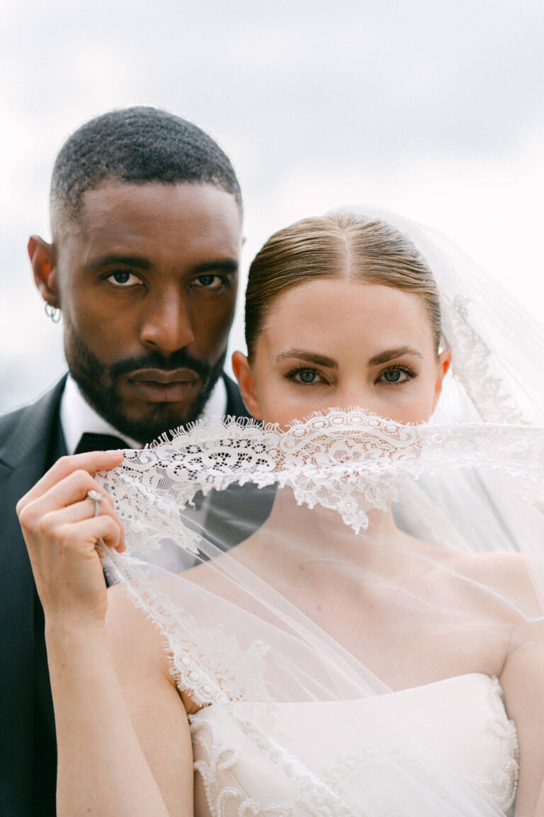 Die Braut im weißen Kleid hält den Spitzenschleier über das Gesicht, der Bräutigam steht dicht dahinter, beide schauen in die Kamera - perfekt für einen Schweizer Hochzeitsplaner, der elegante italienische Hochzeitsinspiration sucht.