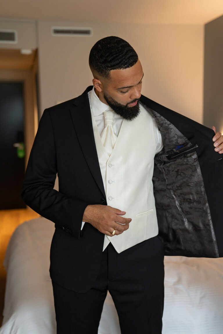 Ein Mann in einem schwarzen Anzug und einer cremefarbenen Weste, möglicherweise ein Wedding Planner, schaut in sein Jackett, während er in einem modernen Raum steht - bereit für eine elegante Hochzeitsplanung in der Schweiz.