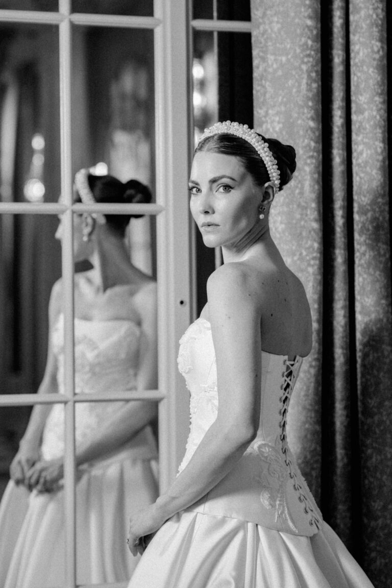 Eine Frau im Hochzeitskleid steht an einem Fenster und schaut über ihre Schulter, ihr Spiegelbild ist im Glas zu sehen - die Eleganz eines Hochzeitsplaners in der Schweiz oder Italien.