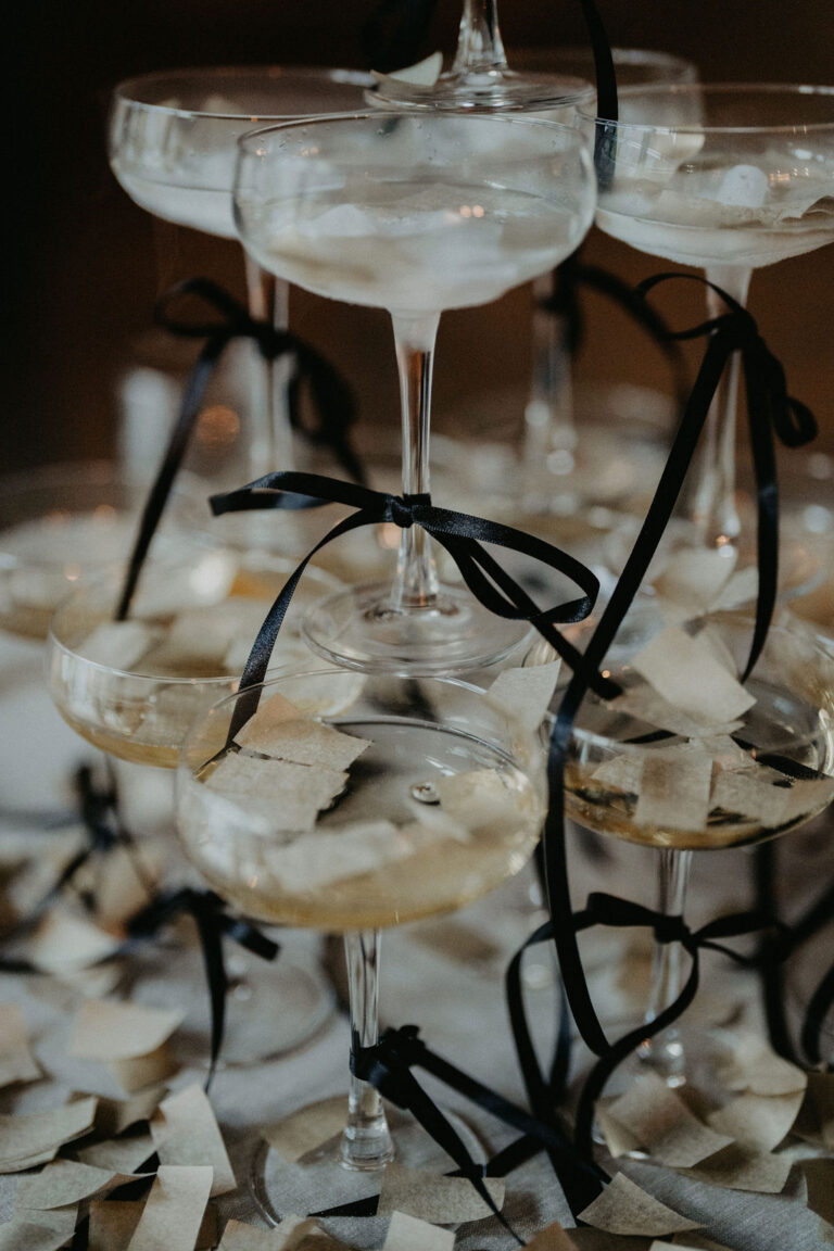 In einem Turm gestapelte Champagnergläser, dekoriert mit schwarzen Bändern und gestreutem Papierkonfetti - ein stilvoller Touch von einem Wedding Planner, inspiriert von eleganten Feiern in der Schweiz und Italien.