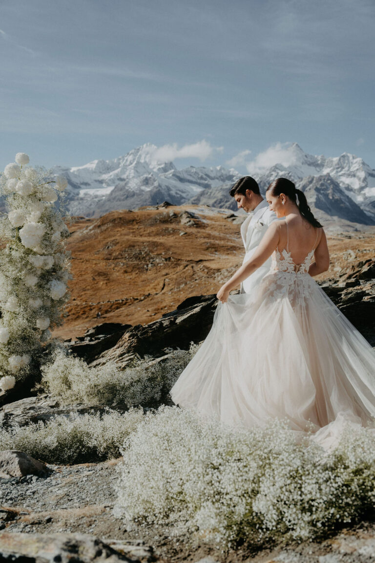 Eine Braut und ein Bräutigam stehen auf einem felsigen Gelände mit Bergen und weißen Blumen im Hintergrund und fangen den Zauber einer Schweizer Hochzeitsplanung inmitten einer atemberaubenden Landschaft ein.