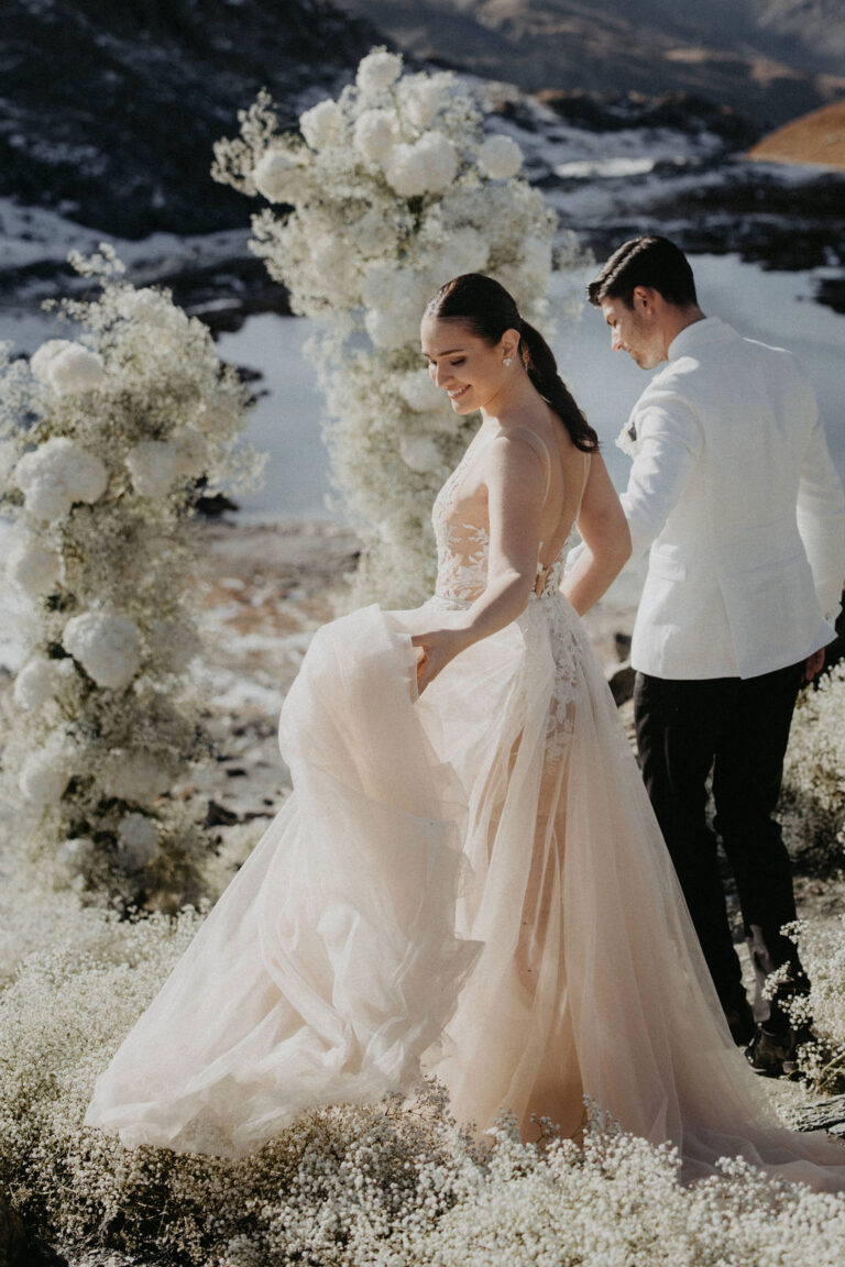 Braut und Bräutigam spazieren im Freien durch weiße Blumenbögen mit Bergen und Wasser im Hintergrund. Ein verträumter Moment, der den Charme der Hochzeitsplanung in der Schweiz zeigt.