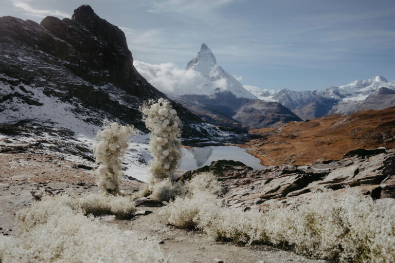 Weiße Blumenarrangements auf felsigem Terrain mit verschneiten Bergen und einem See im Hintergrund unter blauem Himmel - die perfekte Inspiration für Ihre Hochzeitsplanung in der Schweiz.