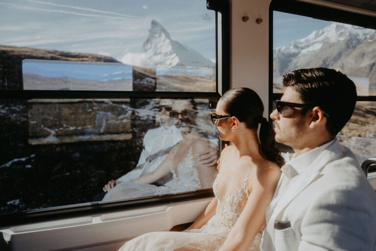 Ein formell gekleidetes Paar sitzt in einem Zug in Italien und blickt auf die Berge, die sich im Fenster spiegeln - vielleicht träumt es mit Hilfe eines örtlichen Wedding Planners von seinem perfekten Tag.