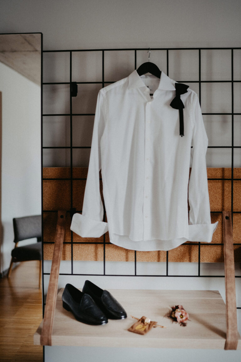 Ein weißes Hemd mit Fliege hängt über schwarzen Schuhen und einer Boutonniere auf einem Holzständer, perfekt arrangiert von einer erfahrenen Hochzeitsplanerin für eine zeitlose Hochzeit in Italien.