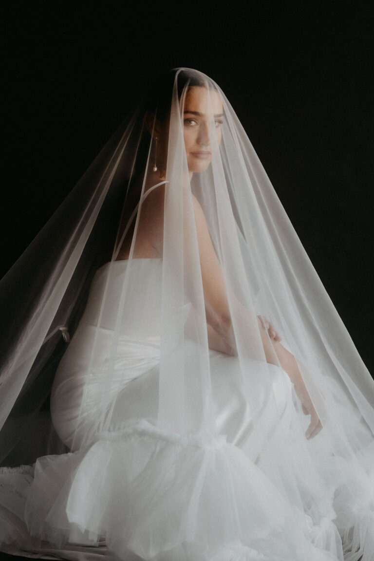 Eine Braut in einem weißen Kleid und langem Schleier sitzt anmutig vor einem dunklen Hintergrund und fängt die Eleganz der Hochzeitsplanung in Italien ein.