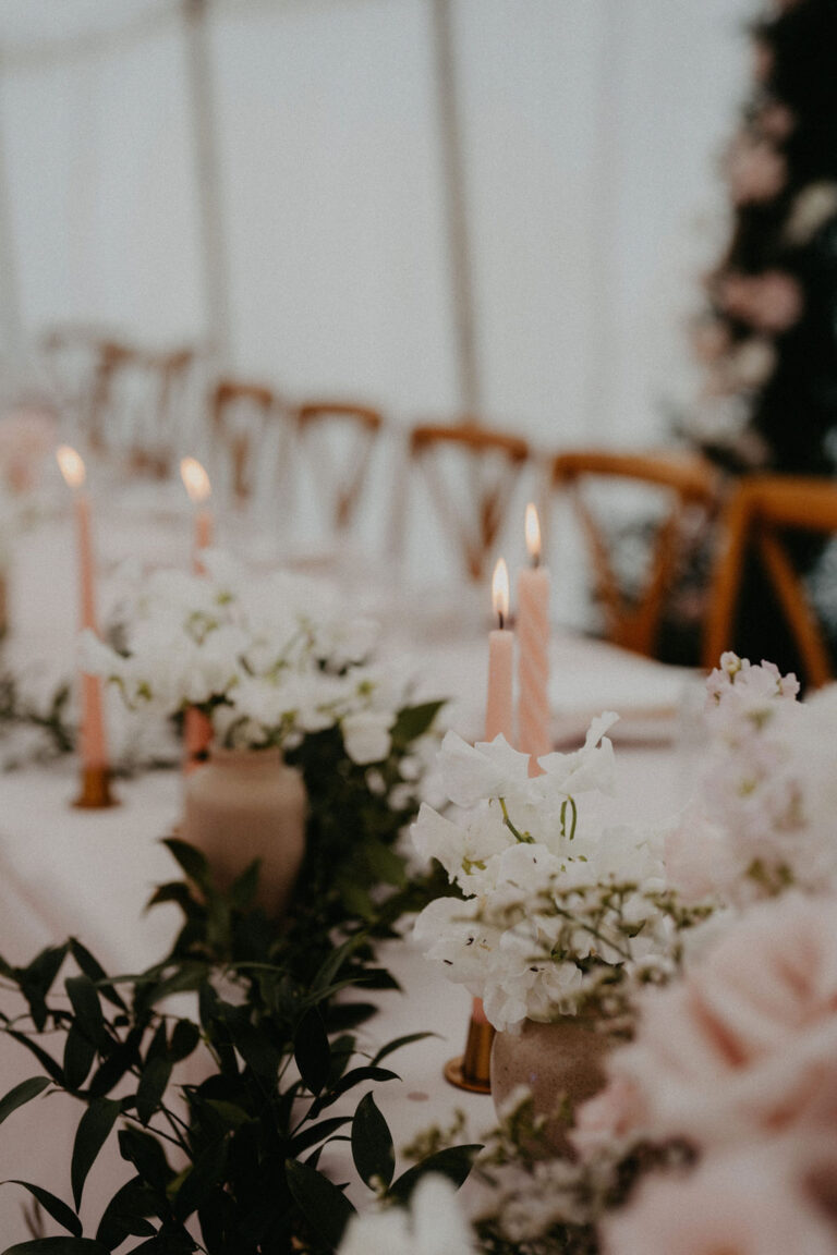 Rosafarbene Kerzen und weisse Blumen schmücken einen elegant gedeckten Tisch für eine Veranstaltung, perfekt für die Hochzeitsplanung mit einem Wedding Planner in der Schweiz, mit Holzstühlen im Hintergrund.