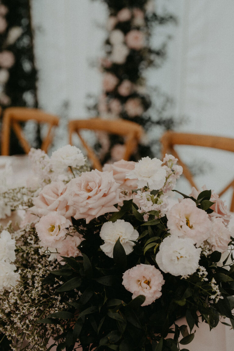 Ein Strauß aus zartrosa und weißen Blumen mit Grünzeug, vor Holzstühlen und Blumendekoration - die perfekte Inspiration für einen Hochzeitsplaner in der Schweiz oder Italien.