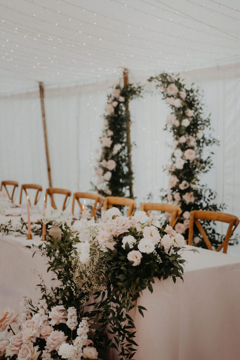 Elegante Hochzeitstafel mit Blumenarrangements, Grünzeug und Holzstühlen unter glitzernden Lichterketten - perfekt gestylt von einem Wedding Planner für Feiern in Italien oder der Schweiz.