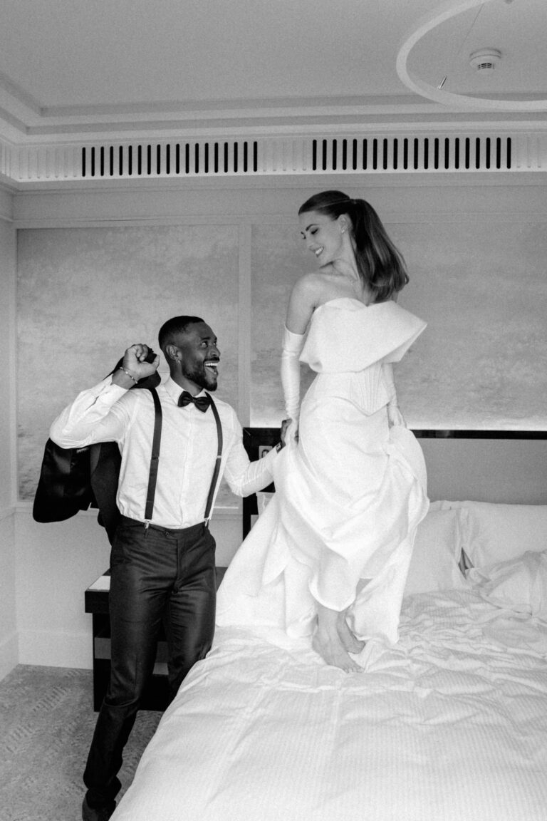 Ein fröhliches Paar in Hochzeitskleidung lacht und hält sich an den Händen, während es auf einem Bett in einem Haus steht. Dies fängt die fröhliche Stimmung einer italienischen und schweizerischen Hochzeit ein - die perfekte Inspiration für jeden Hochzeitsplaner.