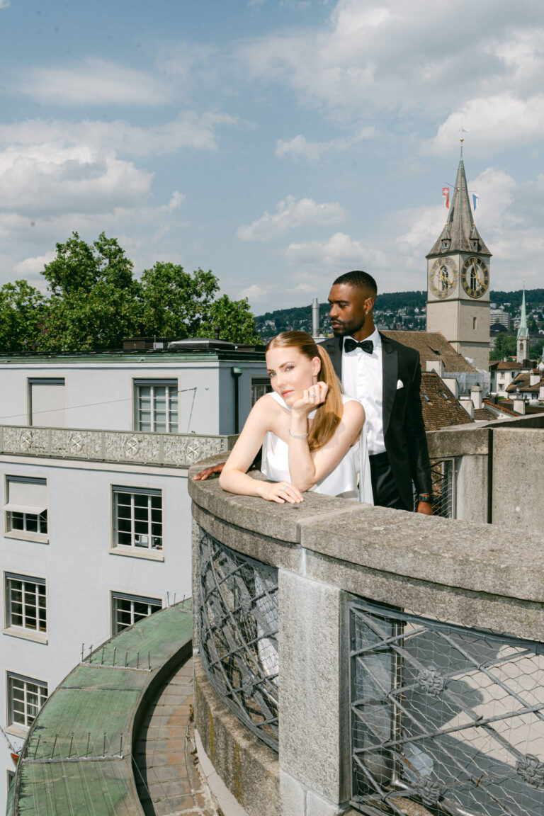 Ein formell gekleidetes Paar steht auf einem Balkon mit Blick auf die Gebäude der Stadt und den Uhrenturm, während ihr Hochzeitsplaner dafür sorgt, dass jedes Detail ihres besonderen Tages in der Schweiz wie aus dem Bilderbuch ist.