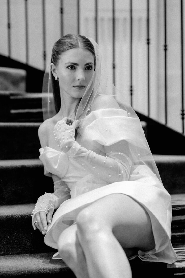 Eine Frau in einem kurzen, schulterfreien Hochzeitskleid mit Schleier sitzt auf einer Treppe und schaut nachdenklich zur Seite - vielleicht träumt sie mit Hilfe eines Hochzeitsplaners von ihrem perfekten Tag.