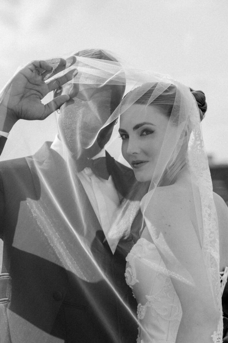 Eine Braut und ein Bräutigam lächeln unter einem durchsichtigen Schleier an ihrem Hochzeitstag in der Schweiz und posieren eng beieinander, wunderschön eingefangen von ihrem Wedding Planner.