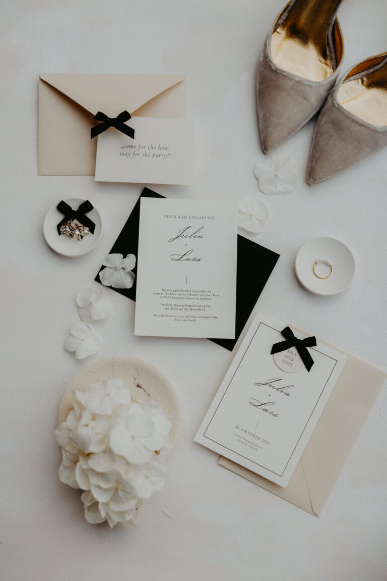 Flache Anordnung von Hochzeitseinladungen, Umschlägen, Ringen, weißen Blütenblättern und beigen Schuhen auf einer weißen Fläche - eine perfekte Inspiration für jeden Hochzeitsplaner in der Schweiz.