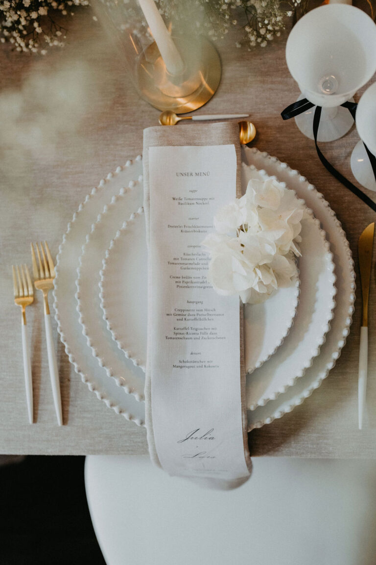 Elegant gedeckter Tisch mit weissen Tellern, goldenem Besteck, einer Menü-Serviette und einer weissen Blumendekoration - eine ideale Inspiration für die Hochzeitsplanung in der Schweiz.