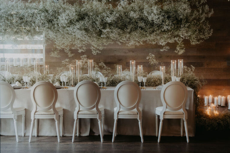 Ein eleganter Tisch mit weißen Stühlen, Kerzen und einem üppigen Blumenarrangement auf einem hölzernen Hintergrund - die perfekte Inspiration für jeden Schweizer oder italienischen Hochzeitsplaner.