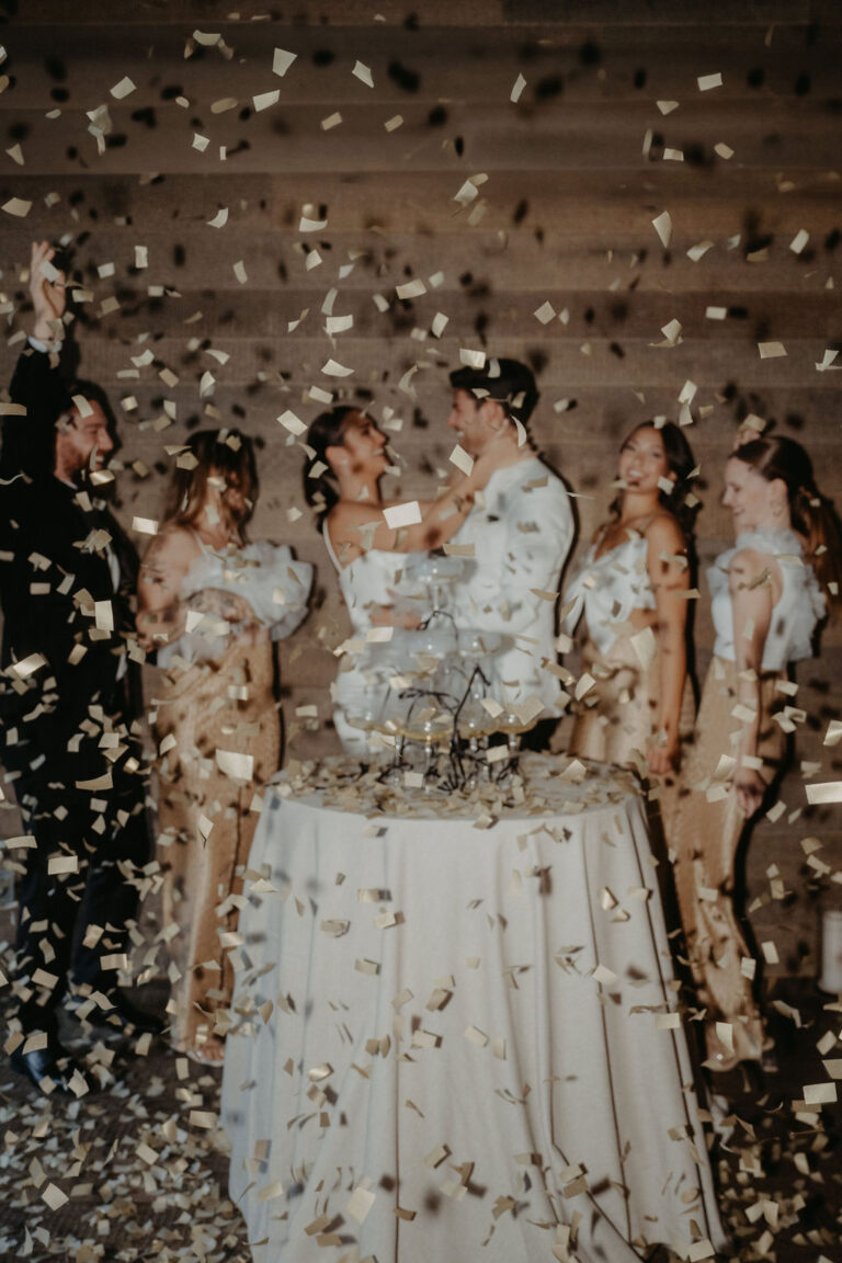 Sechs Personen feiern um eine Torte herum, während goldenes Konfetti fällt und eine fröhliche und festliche Atmosphäre schafft - die perfekte Inspiration für alle Hochzeitsplaner, die Ideen für unvergessliche Hochzeitsfeiern suchen.