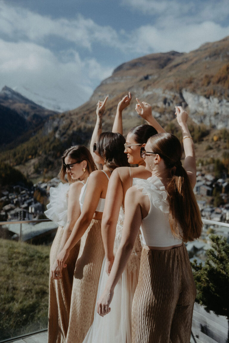 Vier Frauen in aufeinander abgestimmten Outfits stehen im Freien, heben die Hände und genießen die Aussicht auf die Berge - die perfekte Inspiration für jeden Wedding Planner, der auf der Suche nach malerischen Orten in der Schweiz oder in Italien ist.