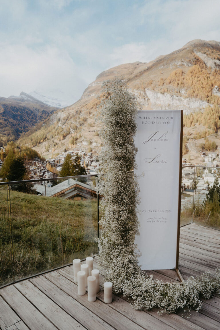 Ein Hochzeitsschild im Freien mit Blumen und Kerzen auf einer Holzterrasse vor der Kulisse der Berge und eines Dorfes in Italien - die perfekte Note für Ihre Hochzeitsplanung.
