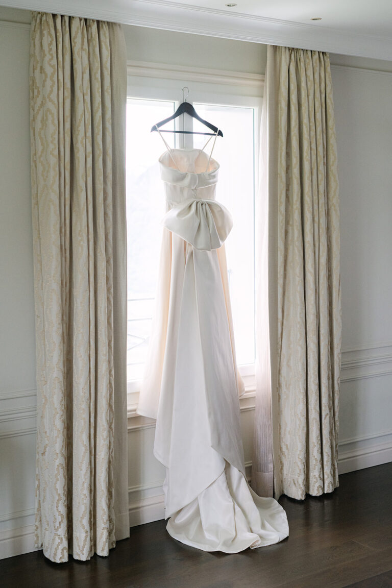 Ein weißes trägerloses Hochzeitskleid hängt an einem Fenster zwischen cremefarbenen Vorhängen in einem hellen Raum.