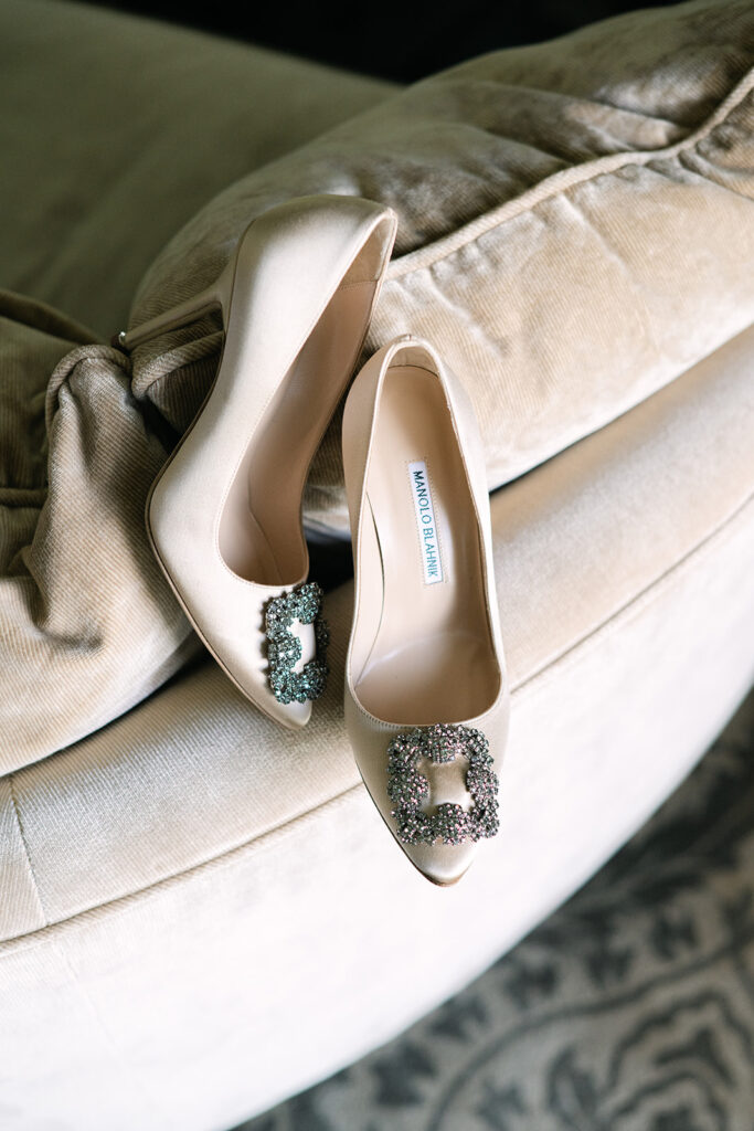 Ein Paar elfenbeinfarbene High Heels mit schmuckvollen Schnallen, perfekt für die Hochzeitsplanung, die elegant auf einem beigen Samtstuhl ruhen. Hier in der Villa Honegg.
