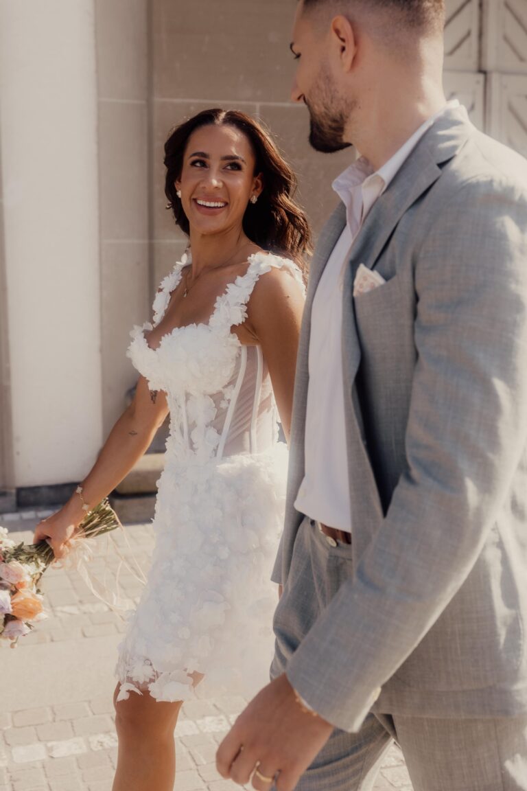 Die Braut im weißen Blumenkleid lächelt einem Mann im hellen Anzug zu, während sie gemeinsam im Freien spazieren gehen. Ihr perfekter italienischer Tag wurde von einem erfahrenen Hochzeitsplaner zum Leben erweckt.