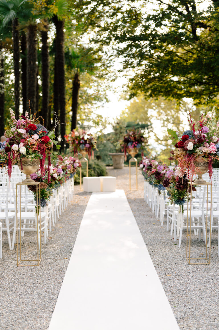 Ein Hochzeitsgang im Freien mit weißen Läufern, eleganten Blumenarrangements und weißen Stühlen in einer von Bäumen gesäumten Umgebung - perfekt kuratiert von einem erfahrenen Hochzeitsplaner für Ihren Traumtag.