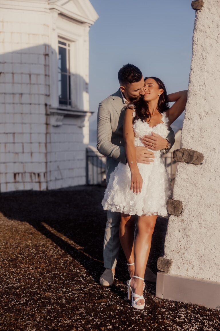 Ein Mann in einem grauen Anzug umarmt und küsst eine Frau in einem weißen Kleid im Freien vor einem weißen Gebäude. Ein zärtlicher Moment, den jeder Wedding Planner in der Schweiz zu schätzen weiß.