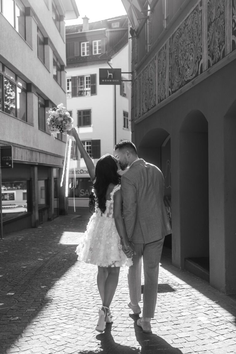 Ein Paar in Hochzeitskleidung geht eine sonnenbeschienene Straße in Italien entlang, hält Hände und einen Blumenstrauß, mit dem Rücken zur Kamera.