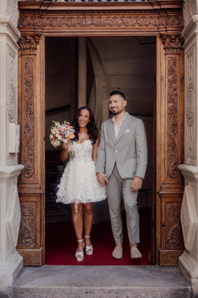Ein Paar in Hochzeitskleidung steht lächelnd in einem verschnörkelten Portal, hält sich an den Händen und hält einen Blumenstrauß - eine inspirierende Szene für die Hochzeitsplanung oder jeden Wedding Planner, der von einer romantischen italienischen Feier träumt.