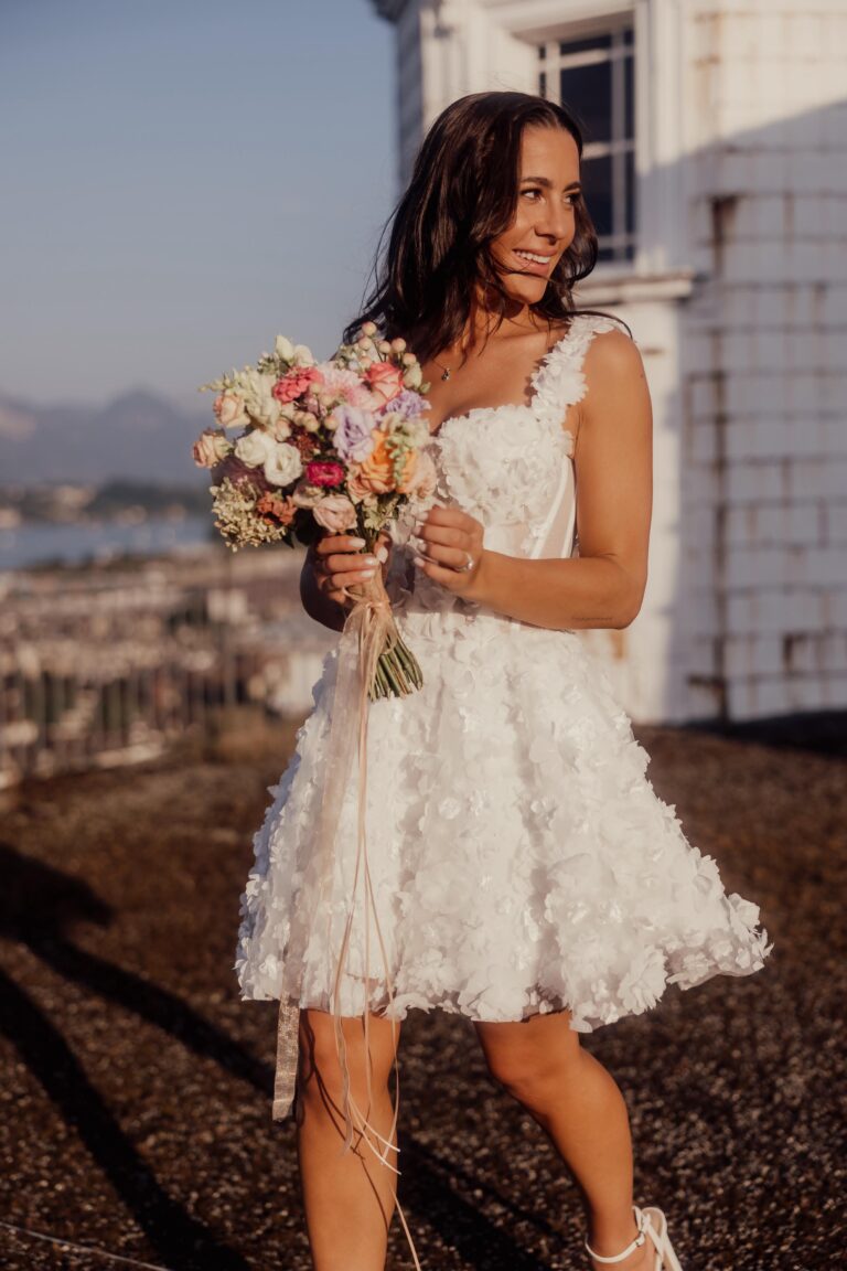 Frau in einem kurzen weißen Blumenkleid mit einem bunten Blumenstrauß in der Hand, lächelnd im Freien im Sonnenlicht - die perfekte Inspiration für einen Schweizer oder italienischen Hochzeitsplaner.