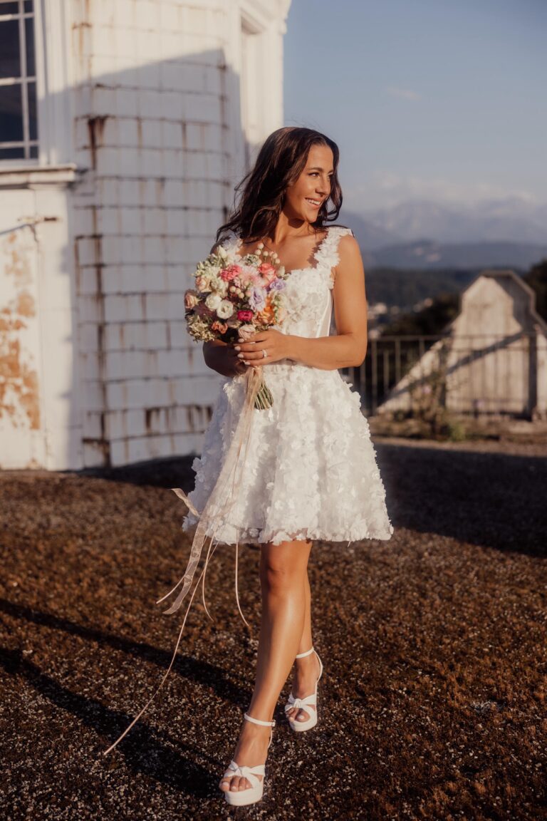 Eine Frau in einem kurzen weißen Kleid, die einen Blumenstrauß hält, lächelt an einem sonnigen Tag im Freien - die perfekte Inspiration für jeden Hochzeitsplaner in der Schweiz.