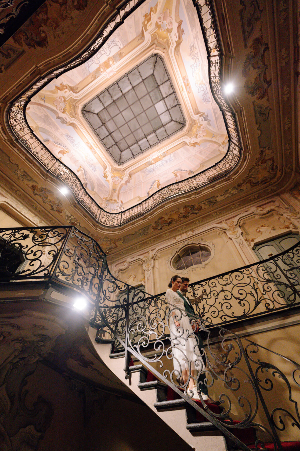 Home 18 Ein Paar steht auf einer prunkvollen Treppe unter einer kunstvoll verzierten Decke und fängt die Eleganz der Hochzeitsplanung in der Schweiz ein.