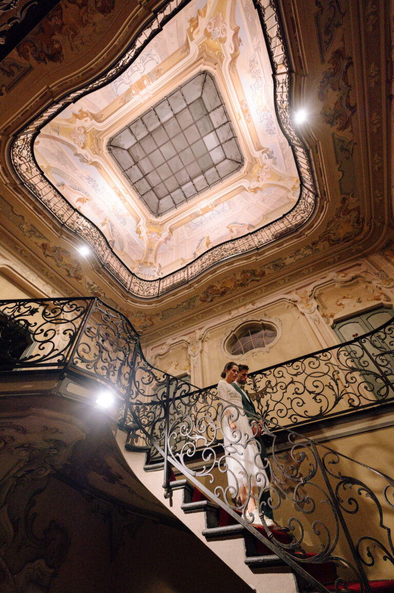 Ein Paar steht auf einer prunkvollen Treppe unter einer kunstvoll verzierten Decke und fängt die Eleganz der Hochzeitsplanung in der Schweiz ein.