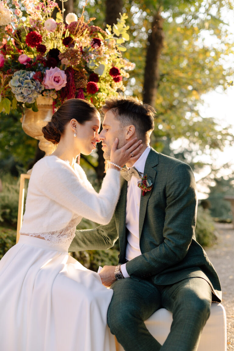Braut und Bräutigam sitzen eng beieinander, berühren sich an den Nasen, umgeben von Blumen im Sonnenlicht - ein bezaubernder Moment, der während ihrer italienischen Hochzeit eingefangen wurde, die von einem erfahrenen Wedding Planner wunderbar koordiniert wurde.