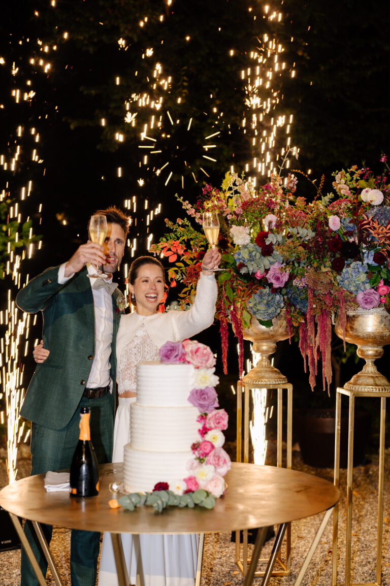 Braut und Bräutigam erheben ihre Sektgläser neben einer gestuften Hochzeitstorte und einem Feuerwerk mit Blumendekoration - ein atemberaubender Moment, inspiriert von der eleganten Hochzeitsplanung in der Schweiz oder Italien.