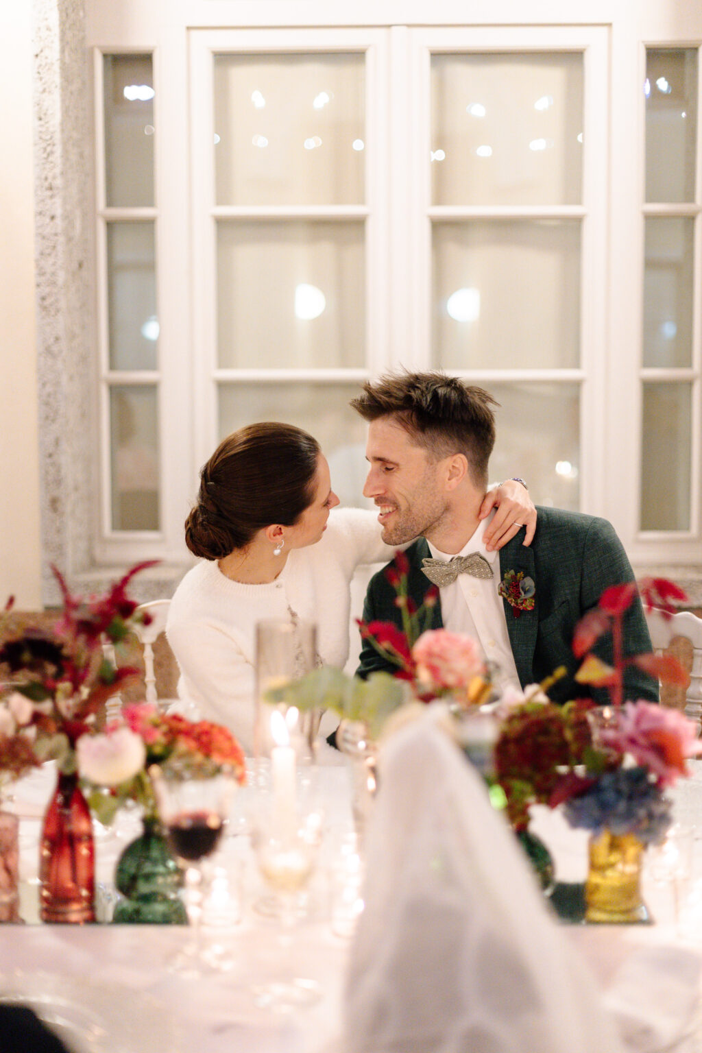 Home 28 Ein Paar sitzt eng beieinander und lächelt sich an einer blumengeschmückten Tafel in weichem, warmem Licht an - die perfekte Inspiration für die Hochzeitsplanung mit dem Wedding Planner Ihres Vertrauens in der Schweiz.
