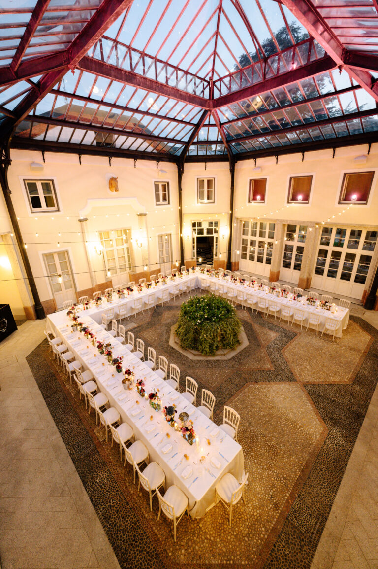 Elegante U-förmige Tische in einem glasüberdachten Innenhof, dekoriert mit Blumen und Lichterketten - die perfekte Inspiration für Ihre Hochzeitsplanung oder zur Weitergabe an Ihren Wedding Planner.