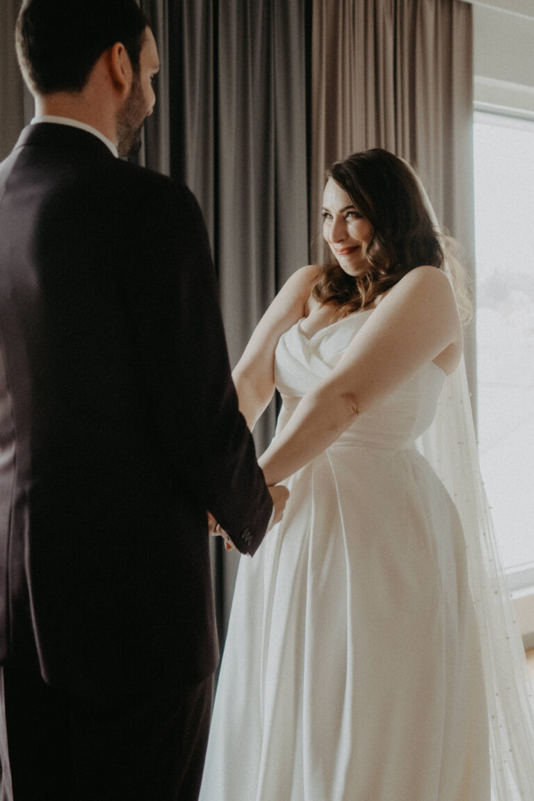 Eine Braut in einem weißen Kleid lächelt ihren Bräutigam an, während sie drinnen an einem Fenster Händchen halten, und fängt damit den Zauber der italienischen Hochzeitsplanung ein.