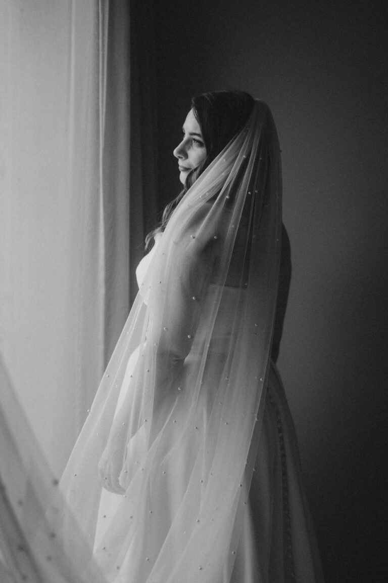 Die verschleierte Braut blickt auf einem Schwarz-Weiß-Foto mit sanftem Seitenlicht aus dem Fenster - ein eleganter Moment, perfekt für jede italienische Hochzeitsplanung.