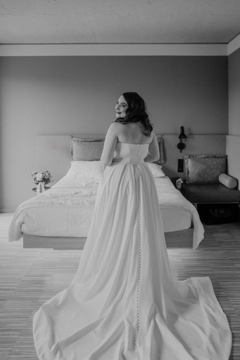 Eine Braut in einem wallenden Kleid steht lächelnd vor einem Bett in einem sanft beleuchteten, modernen Schlafzimmer und fängt die Essenz stilvoller Hochzeitsplanung in der Schweiz ein.