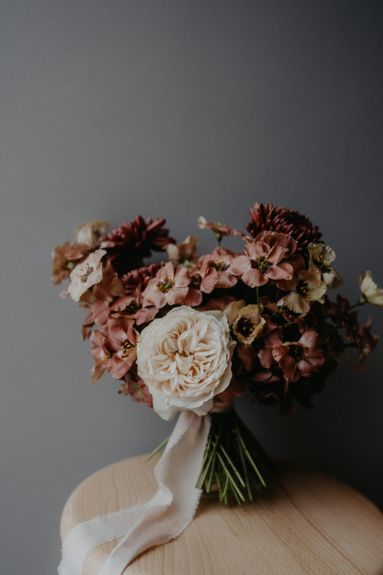 Ein Strauß aus rosafarbenen, burgunderroten und cremefarbenen Blumen mit weißem Band, arrangiert von einem erfahrenen Wedding Planner in Italien, ruht elegant auf einer Holzfläche.