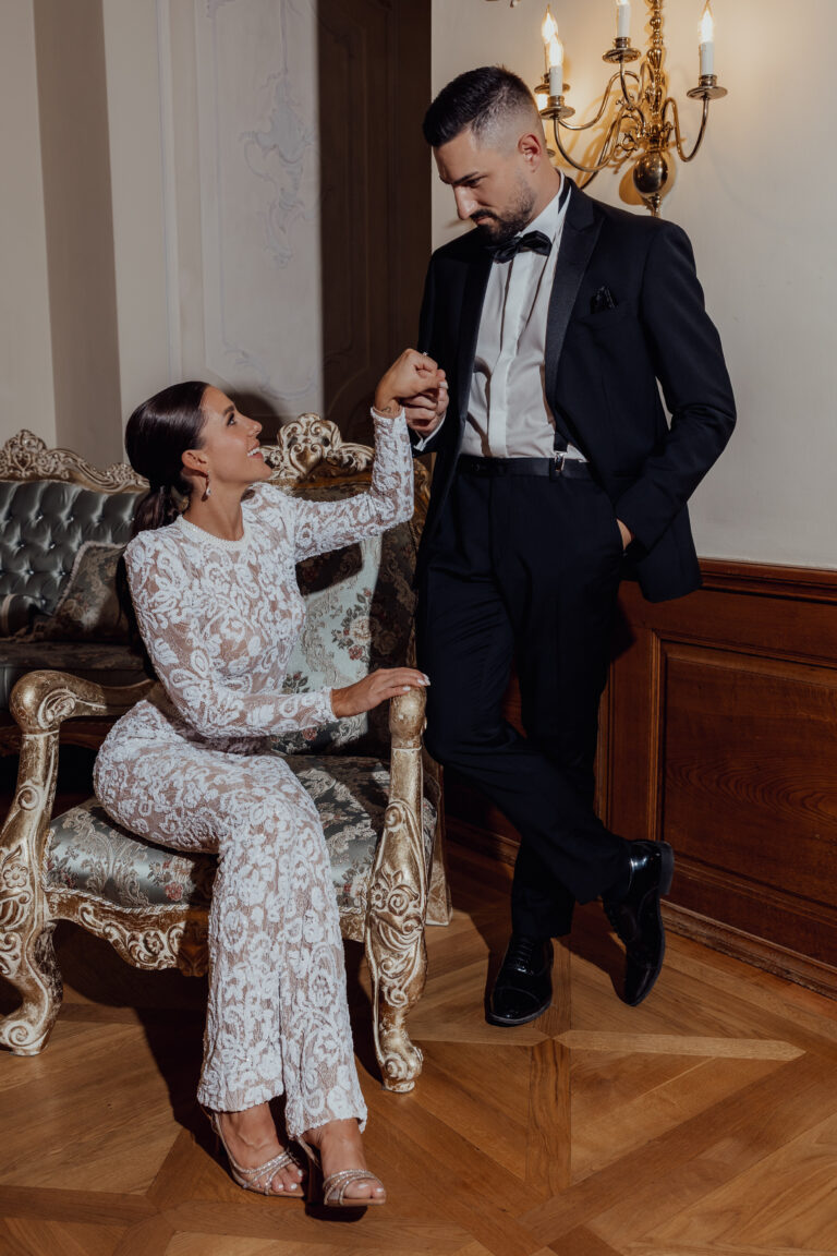 Eine Frau in einem weißen Spitzenoverall sitzt auf einem verzierten Stuhl und hält Händchen mit einem Mann im schwarzen Smoking.