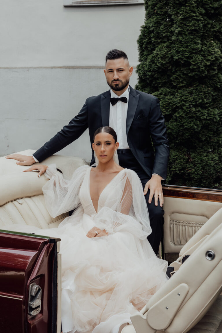 Die Braut in einem weißen Kleid und der Bräutigam in einem schwarzen Smoking posieren in einem Oldtimer-Cabrio im Freien.