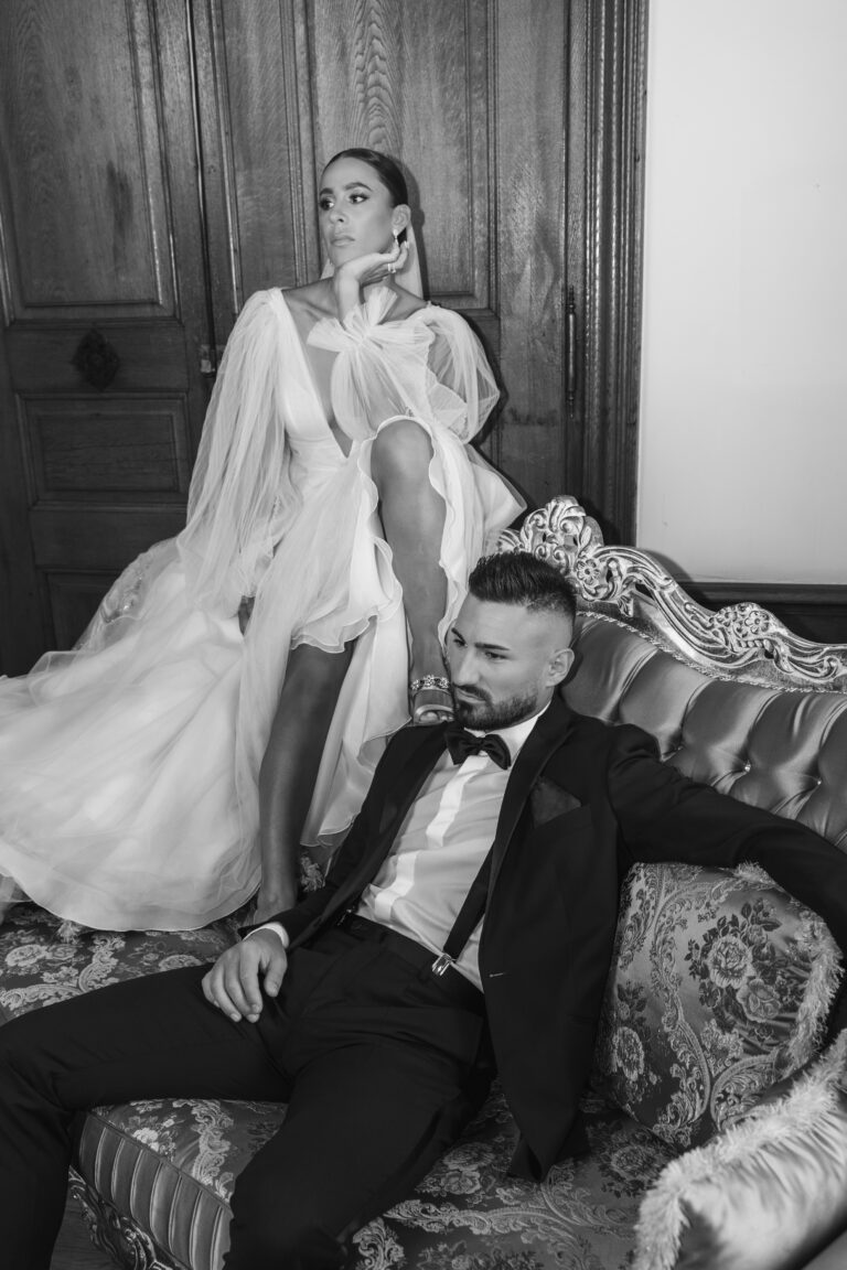 Eine Frau in einem wallenden Kleid steht hinter einem Mann im Anzug, beide posieren auf einem verzierten Sofa in einem stilvollen Raum.